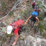 Hikers climbing a rocky, steep section of bush trail together, each carrying backpacks and assisting one another through dense scrub.
