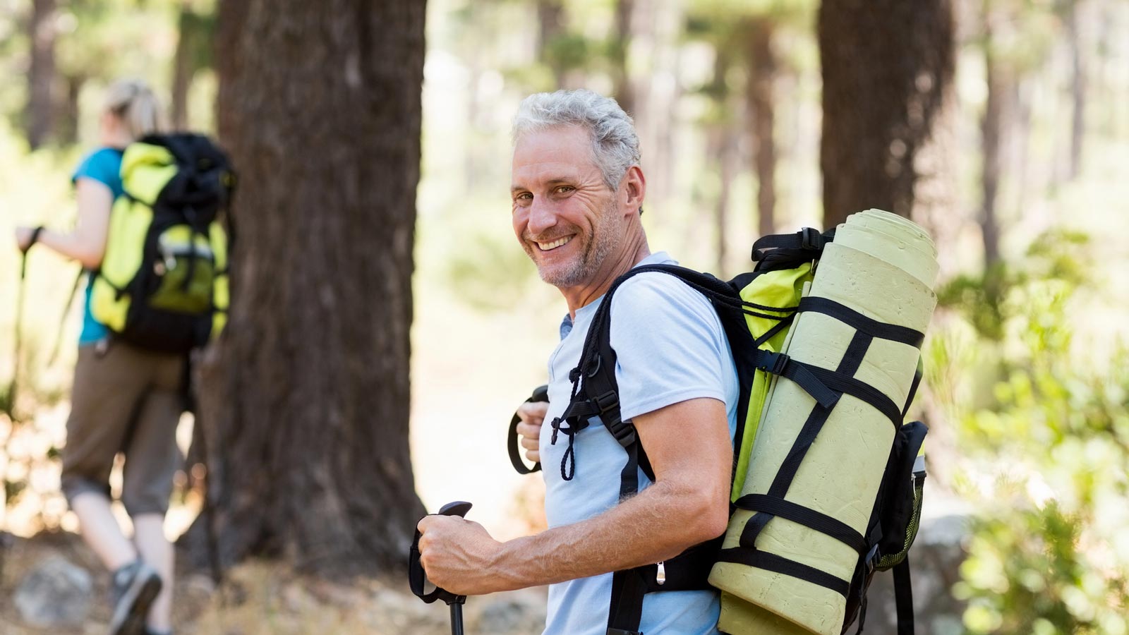 Hiker smiling at the camera while carrying a backpack with a rolled sleeping mat, trekking through an australian forest with another hiker in the background.