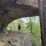 Hiker standing beneath a large rock overhang providing natural shelter from sun and weather