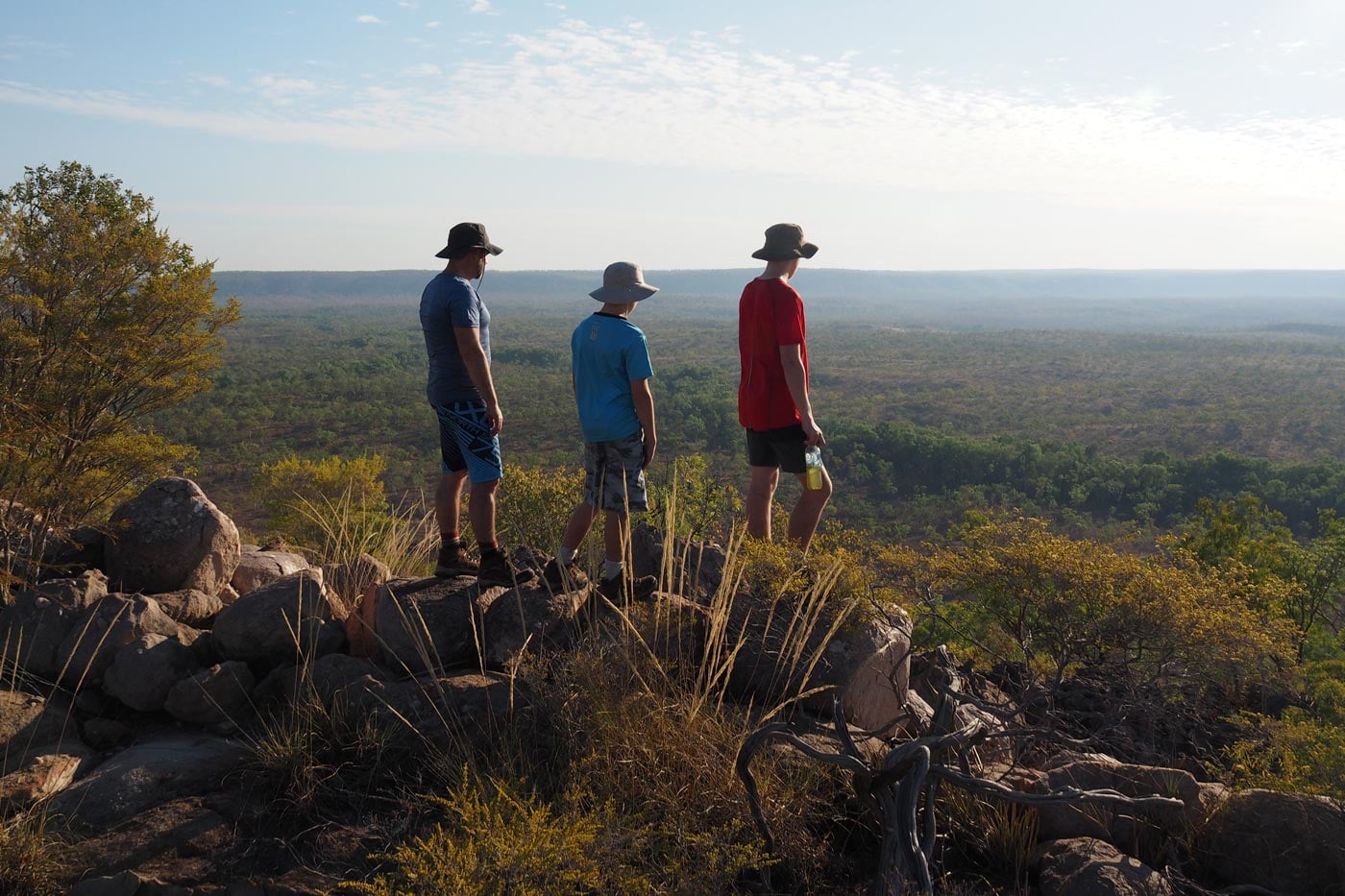 Three hikers stand on a rocky overlook on the jatbula trail, northern territory, gazing out across a vast, sun-drenched landscape of green trees and savanna stretching toward a flat horizon.