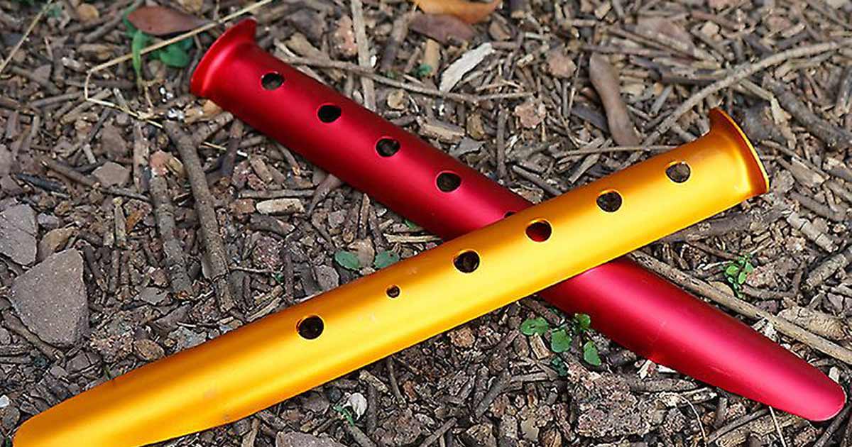 Wide aluminium snow and sand pegs with perforated bodies lying on loose ground, designed for use in soft or sandy terrain.