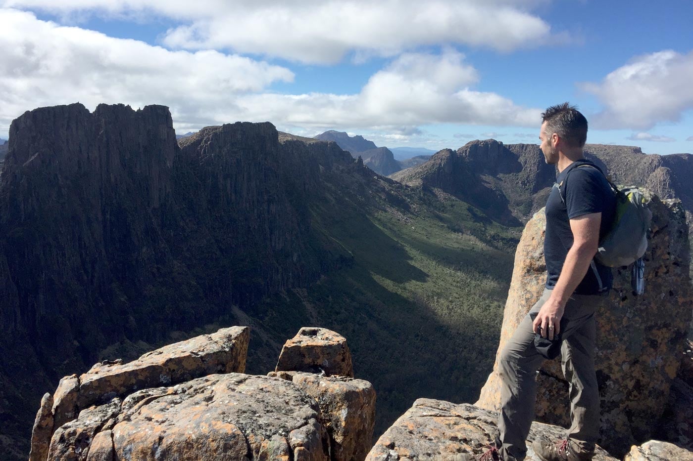 A side profile of a hiker standing on a rugged, rocky outcrop at the summit of the acropolis in tasmania, overlooking a deep, verdant valley and dramatic, jagged mountain peaks in the distance.