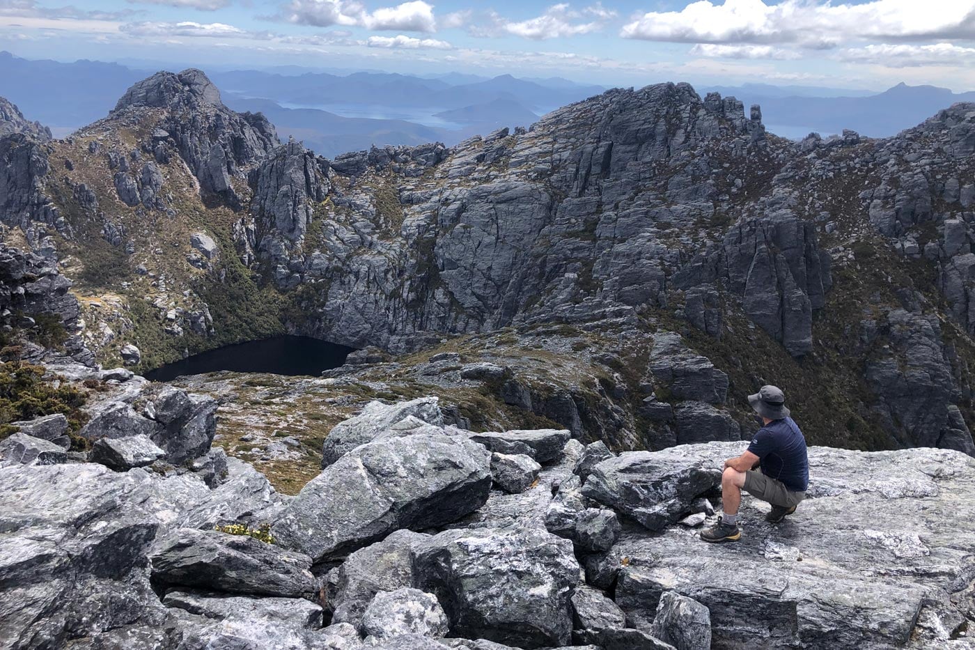 A hiker in a hat and dark t-shirt sits on a jagged, grey rock ledge in the foreground, looking out over a dramatic mountain landscape. Below him, a dark, still alpine lake is cradled by steep, rocky cliffs, while layers of blue mountain ranges and water stretch toward the distant horizon under a cloudy sky.