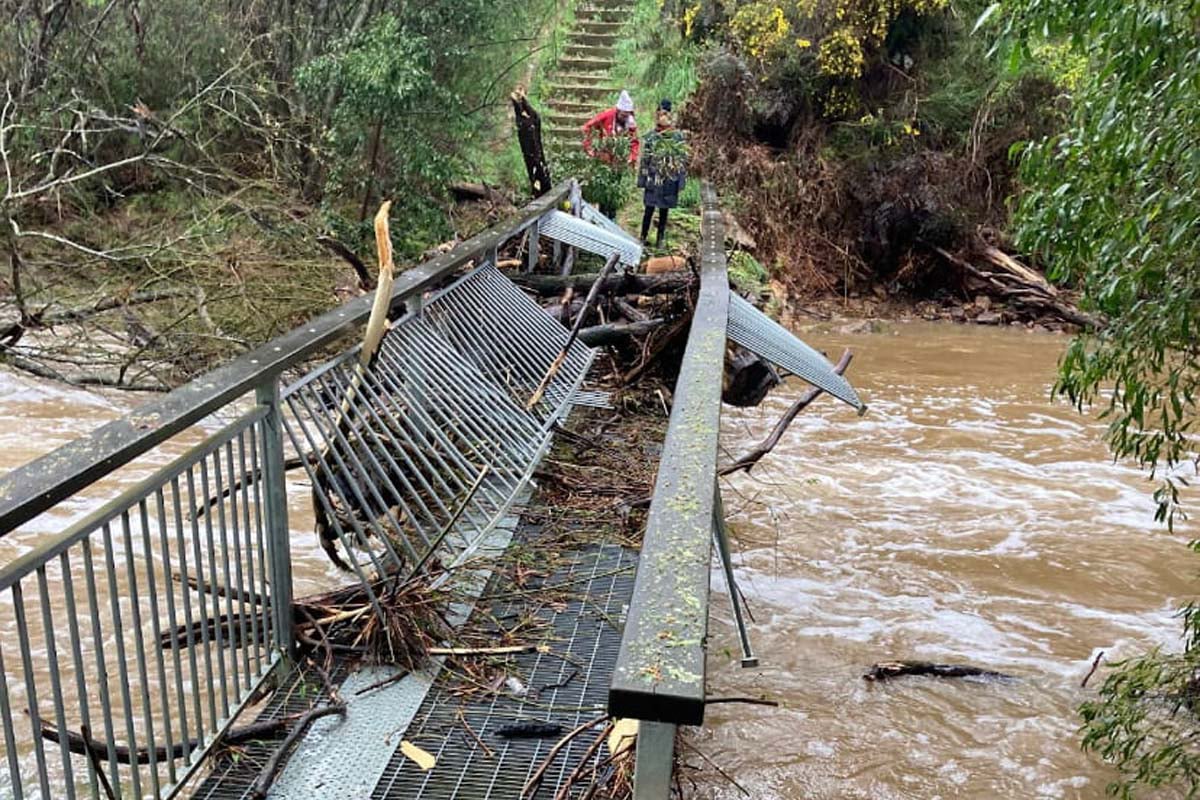 A damaged metal bridge over a flooded river, with debris piled against it, showing the destructive power of flash floods after heavy rain.