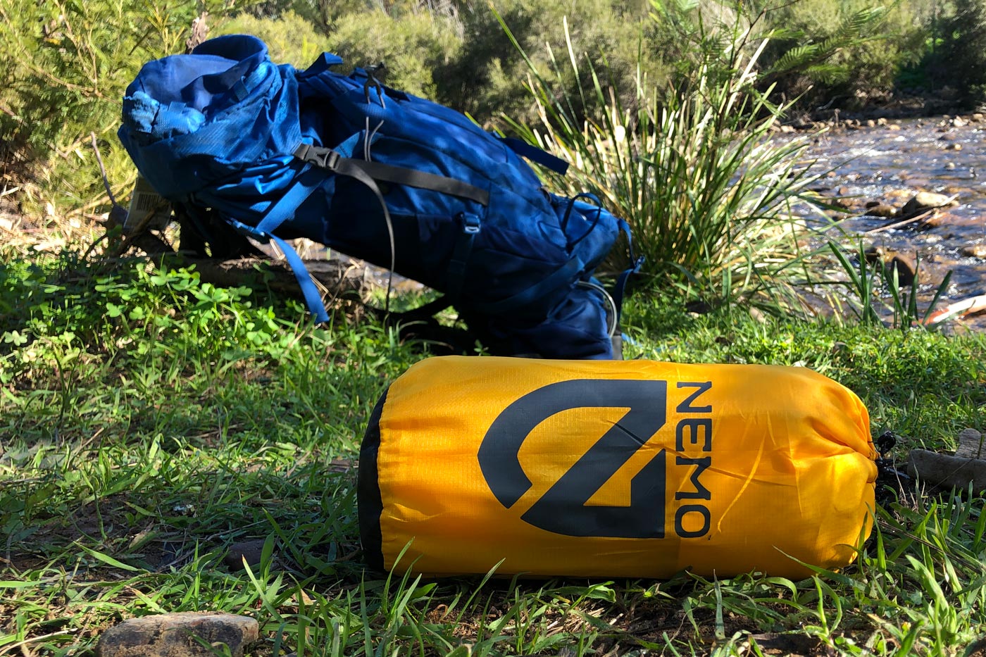 A compacted sleeping mat in a storage sack beside a backpack, ready for packing during a hiking trip.