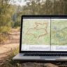 Laptop showing online hiking route planning maps and elevation profiles, placed on a dirt trail in australian bushland with eucalyptus trees in the background.