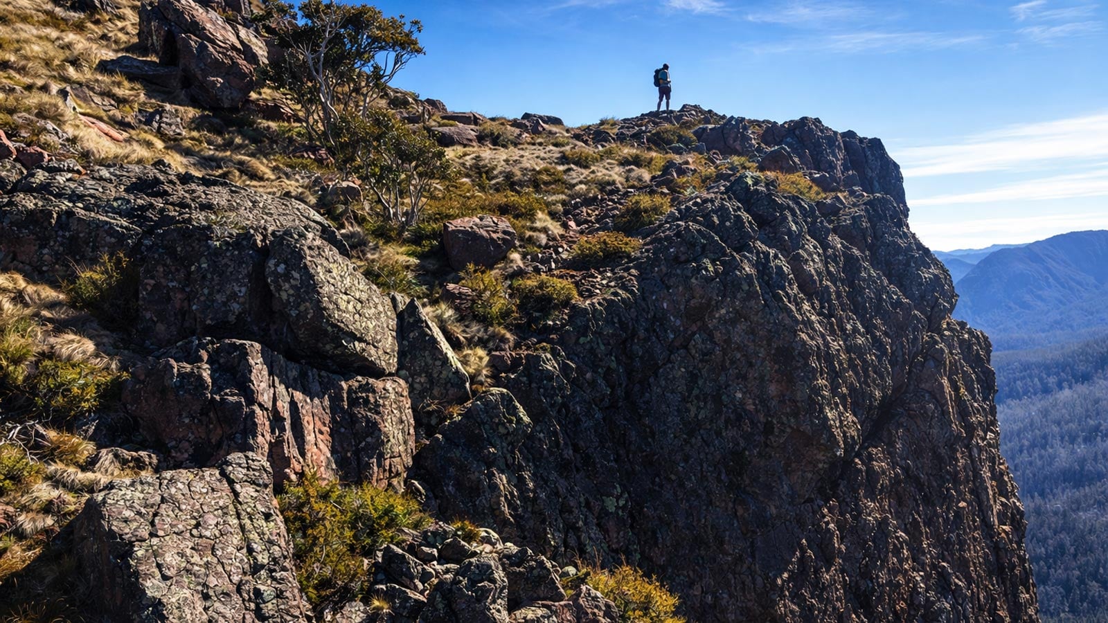 A hiker standing on a rocky trail overlooking a vast landscape, viewed from behind, small in frame, demonstrating scale and human presence in outdoor photography.