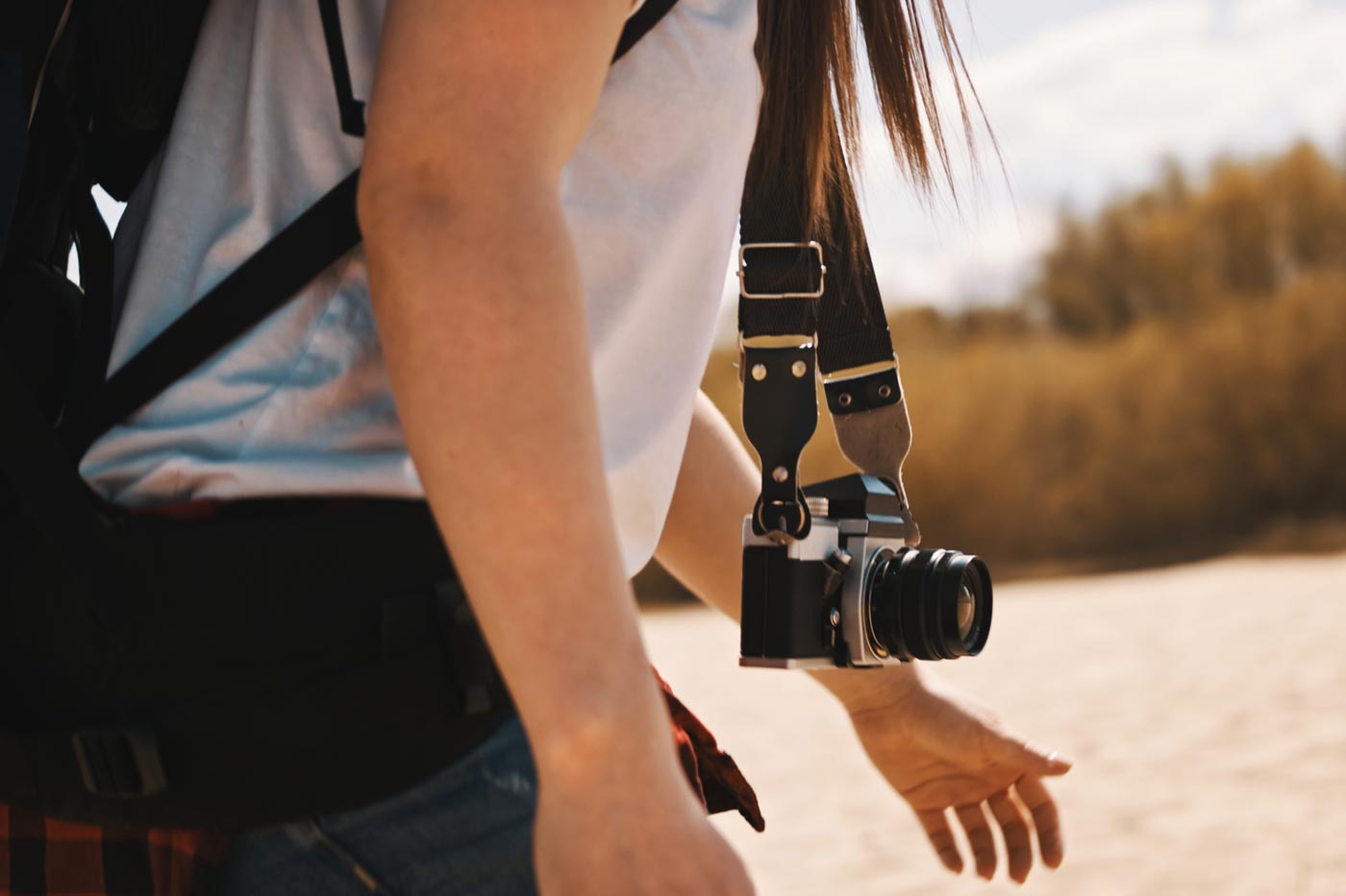 Hiker walking with a camera hanging from a shoulder strap, wearing a backpack and outdoor clothing in a natural outdoor setting.