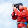 Hiker wearing a red jacket and backpack rain cover using a camera to photograph the landscape outdoors under cloudy skies.