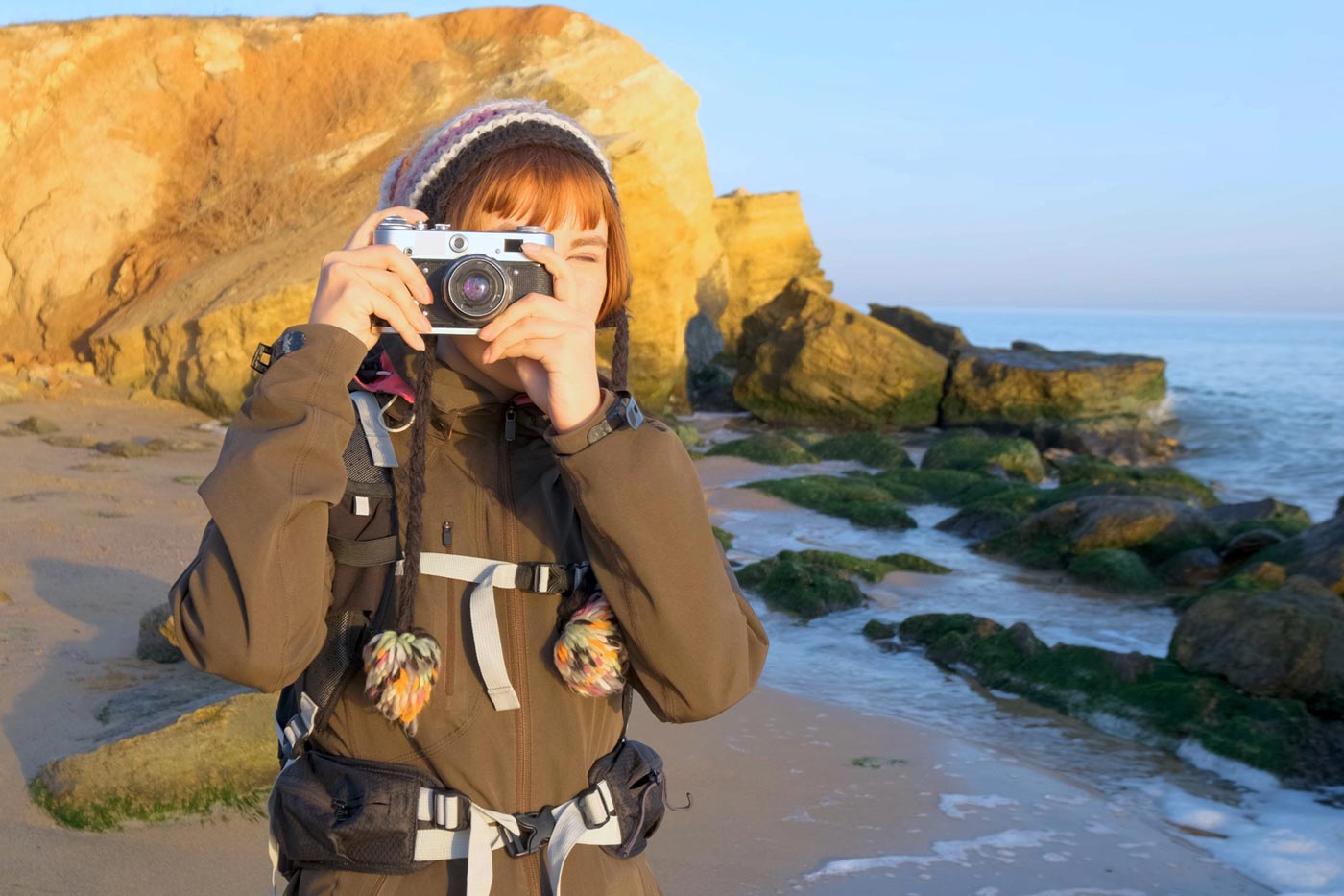 Hiker standing on a rocky beach using a compact camera to take a photo, wearing a backpack and outdoor clothing near the coastline.