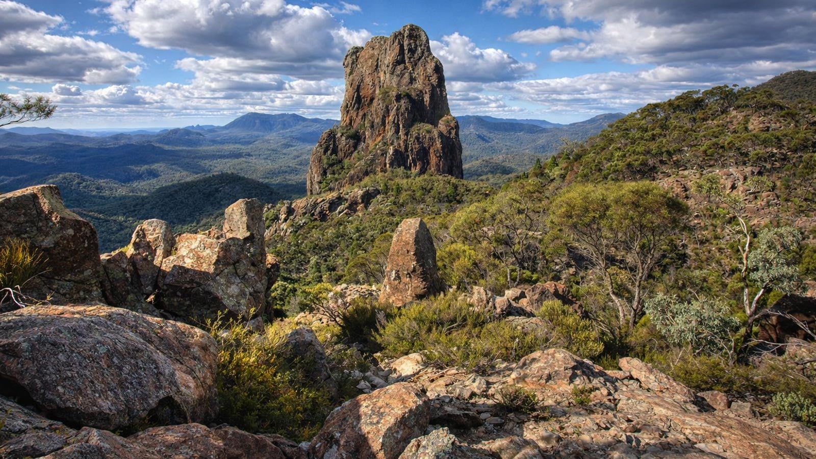 Rocky lookout with scattered boulders and low woodland, looking towards the prominent belougery spire and surrounding ranges in warrumbungle national park.