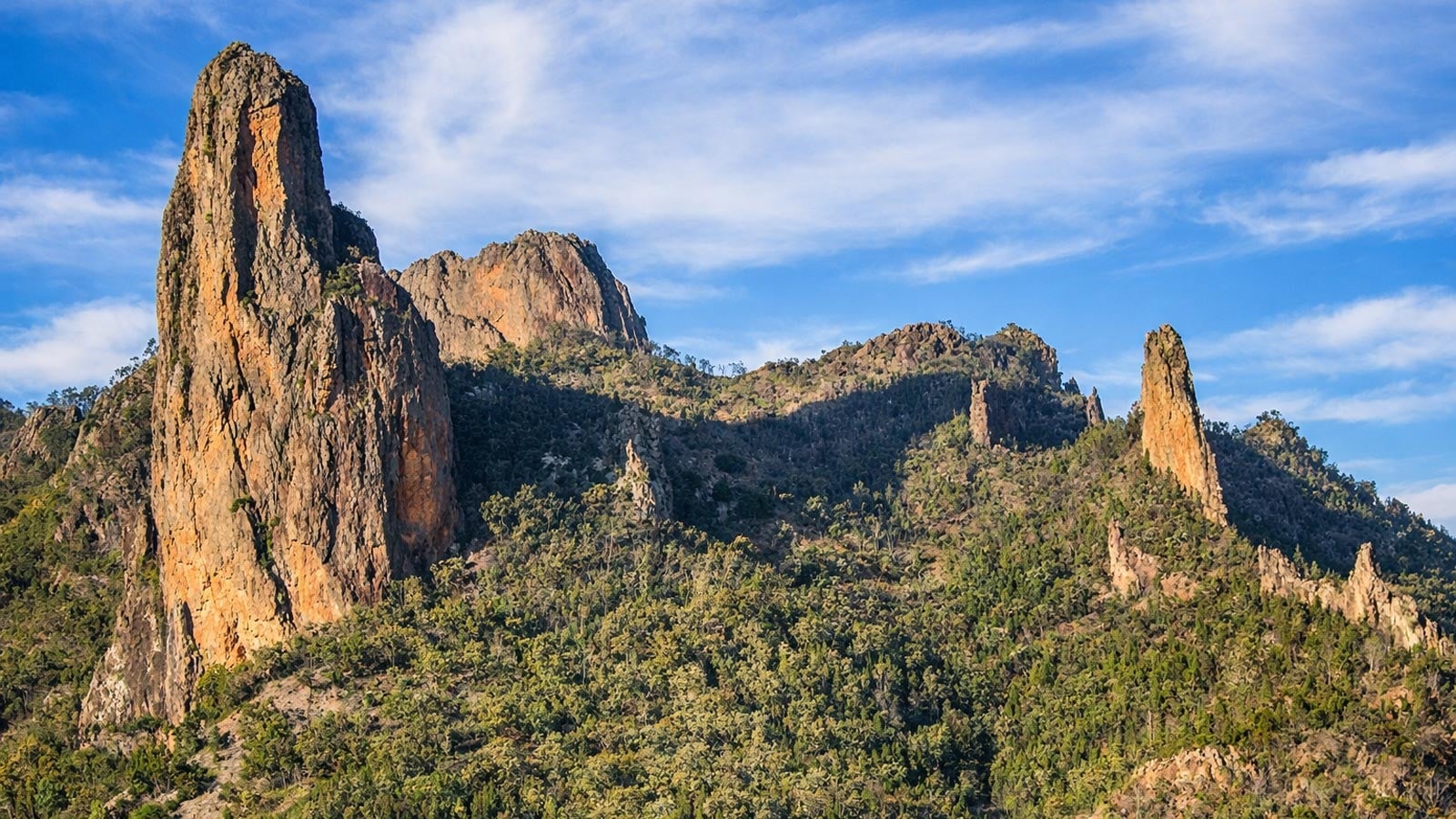Tall volcanic rock spires and rugged cliffs rising above forested slopes in warrumbungle national park under a blue sky.