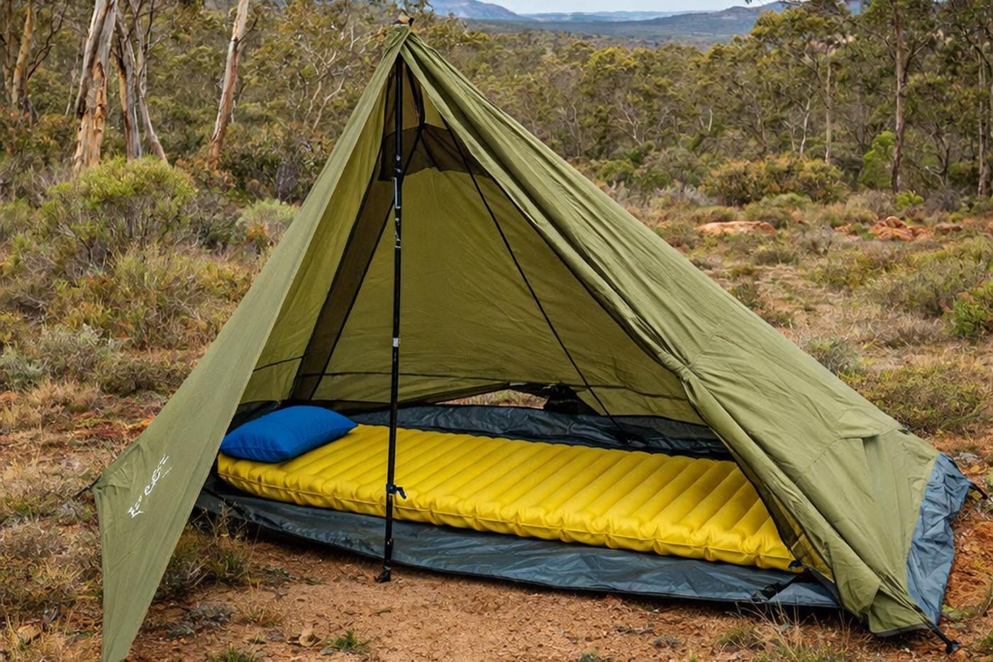 Hiking pole tent pitched with trekking poles in open bushland, showing a single-wall shelter with sleeping mat inside.