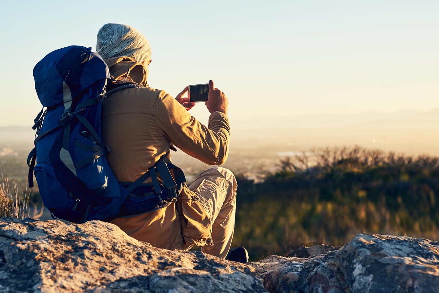 Hiker sitting on rocky ground using a smartphone to photograph the landscape, wearing a backpack and outdoor clothing in natural light.