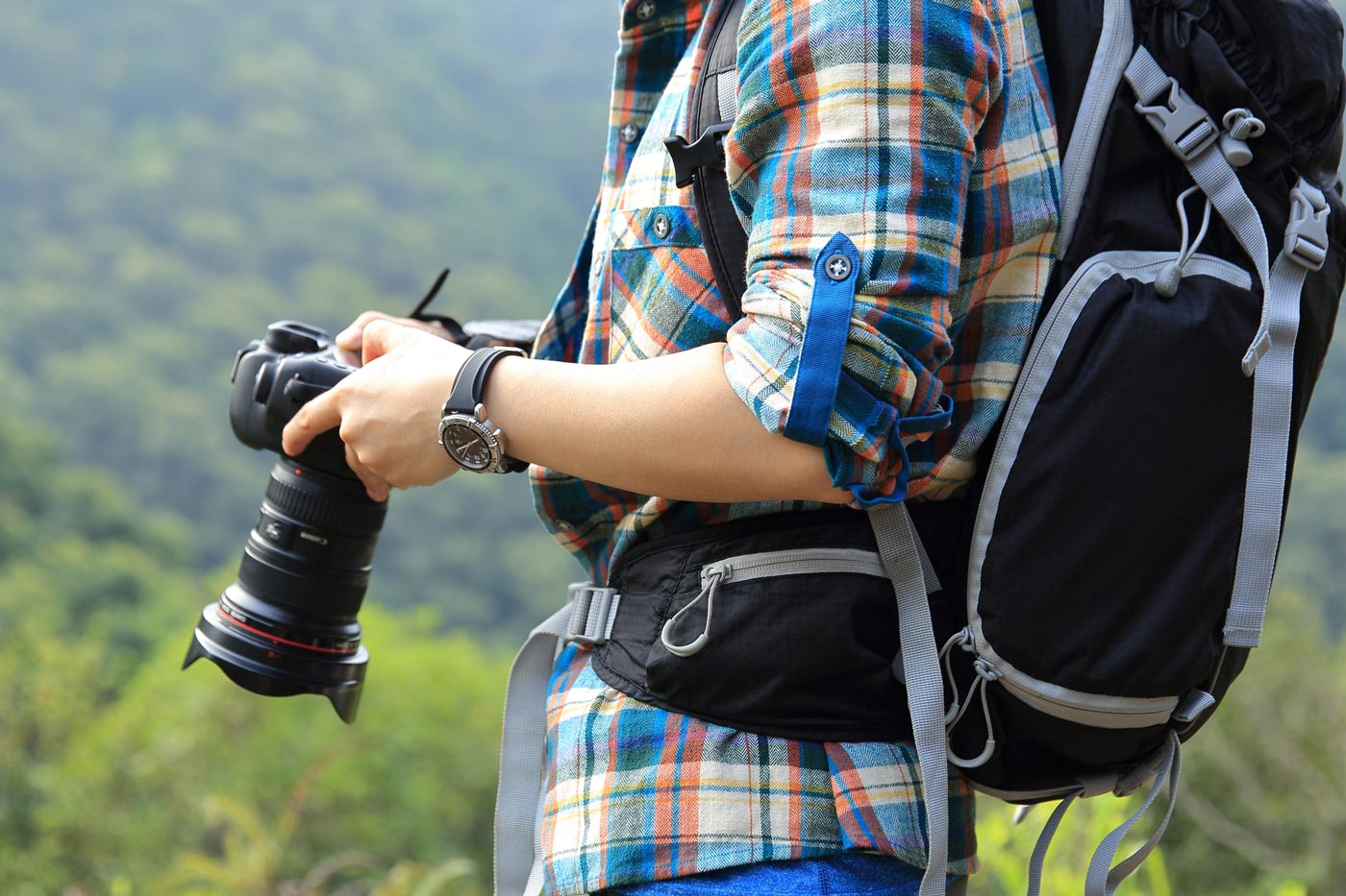 Hiker wearing a backpack holding an interchangeable-lens camera while standing outdoors, with forested hills in the background.