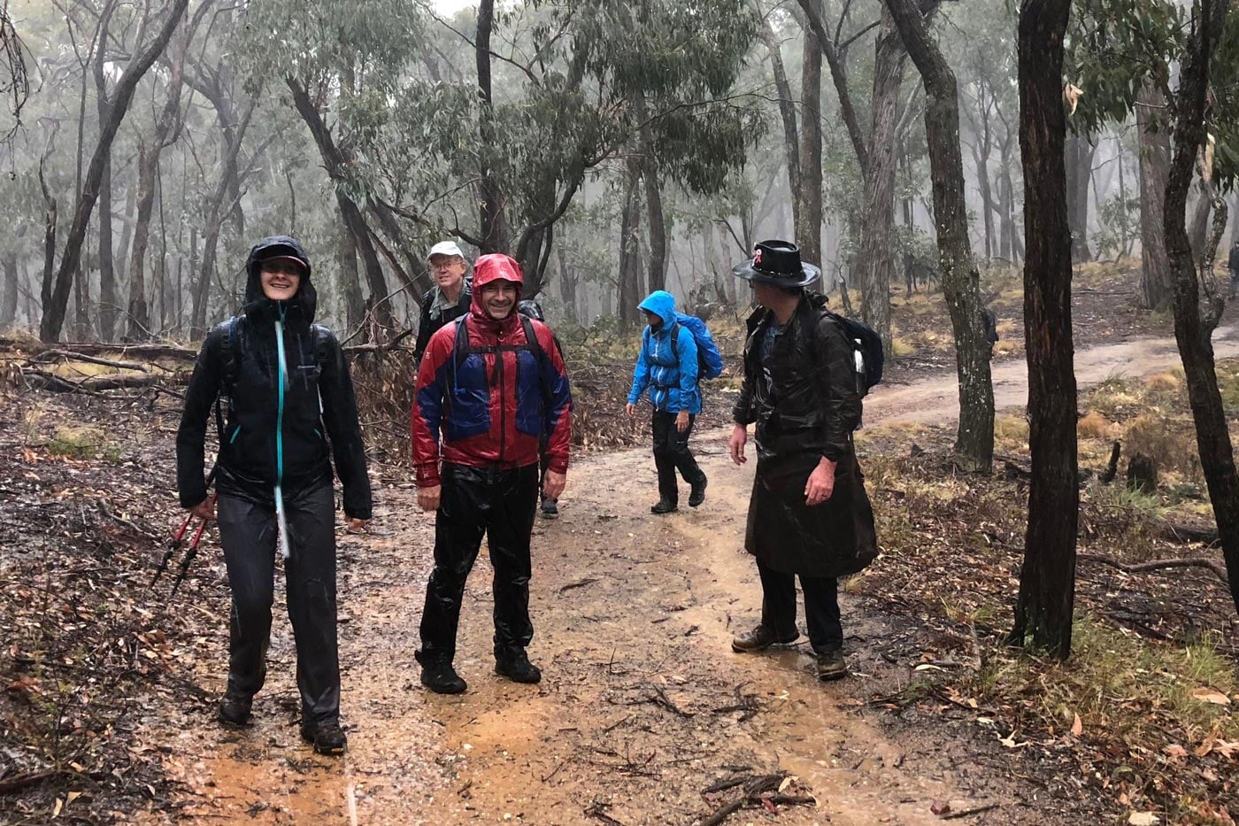 Group of hikers walking along a muddy forest track in heavy rain, wearing wet-weather jackets and packs, illustrating how prolonged moisture challenges breathable and waterproof hiking gear