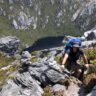 Hiker ascending steep rocky alpine terrain in tasmania carrying a loaded overnight backpack above a mountain lake