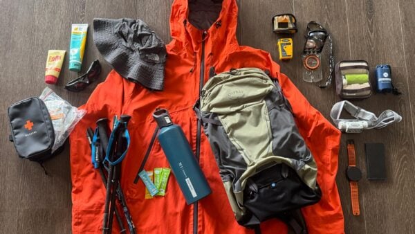 Gear-for-a-day-hike Day hiking gear laid out on a table including backpack, rain jacket, first aid kit, map, compass, trekking poles, water bottle, headlamp, emergency blanket and other safety essentials.