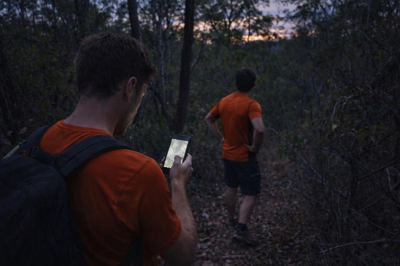 Two hikers off track in dense bush at dusk, one checking a phone for navigation