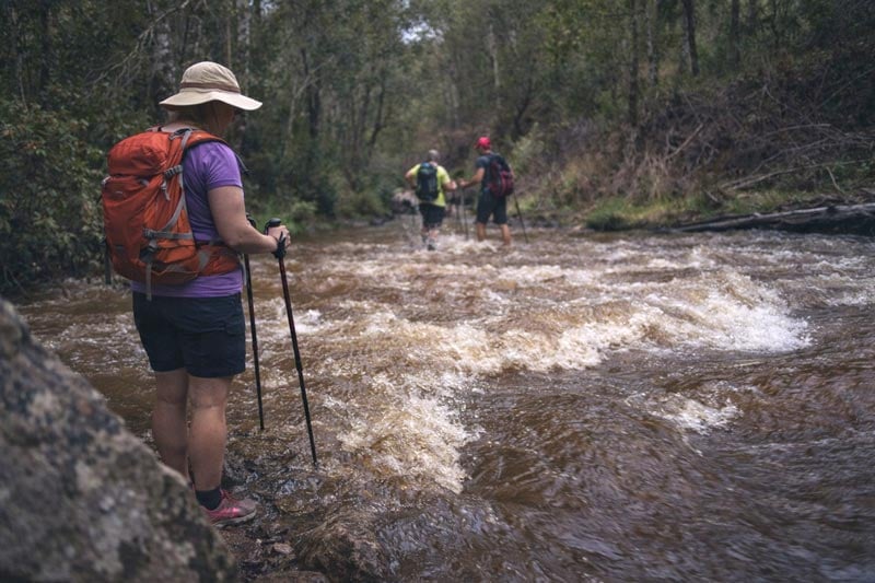 Hiker pauses at the edge of a fast-flowing river while others begin crossing ahead