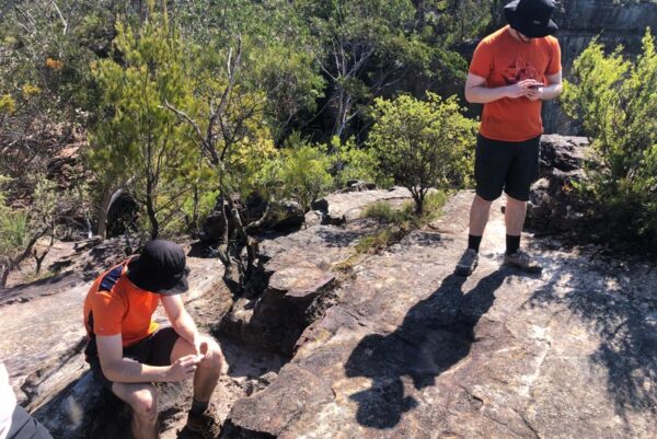 Hikers paused on a bush track in the blue mountains, with one resting while others check navigation, highlighting a group decision point as pace differences emerge