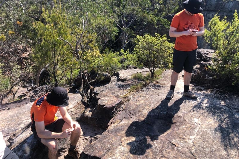Hikers paused on a bush track in the blue mountains, with one resting while others check navigation, highlighting a group decision point as pace differences emerge