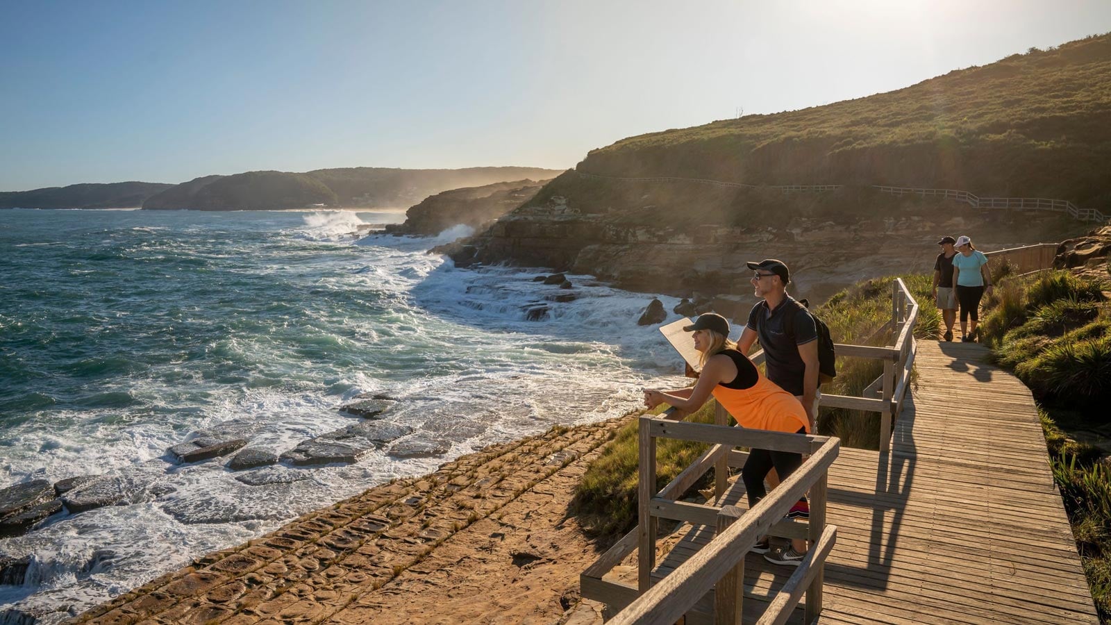 The Hiker's Guide to Whale Watching in NSW Hikers pausing on the bouddi coastal walk boardwalk to look out over crashing waves and sandstone rock platforms
