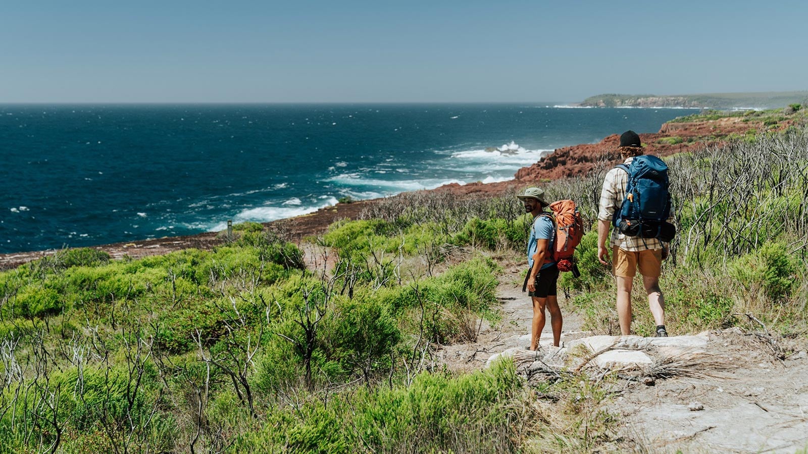 The Hiker's Guide to Whale Watching in NSW Two hikers with backpacks pausing on the light to light walk with red rock headlands and wild ocean behind, beowa national park