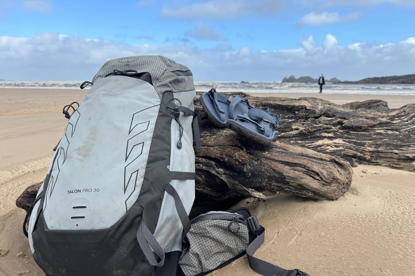 A grey technical daypack sits on the sand next to a piece of driftwood holding a pair of zorali trailblazer sandals.