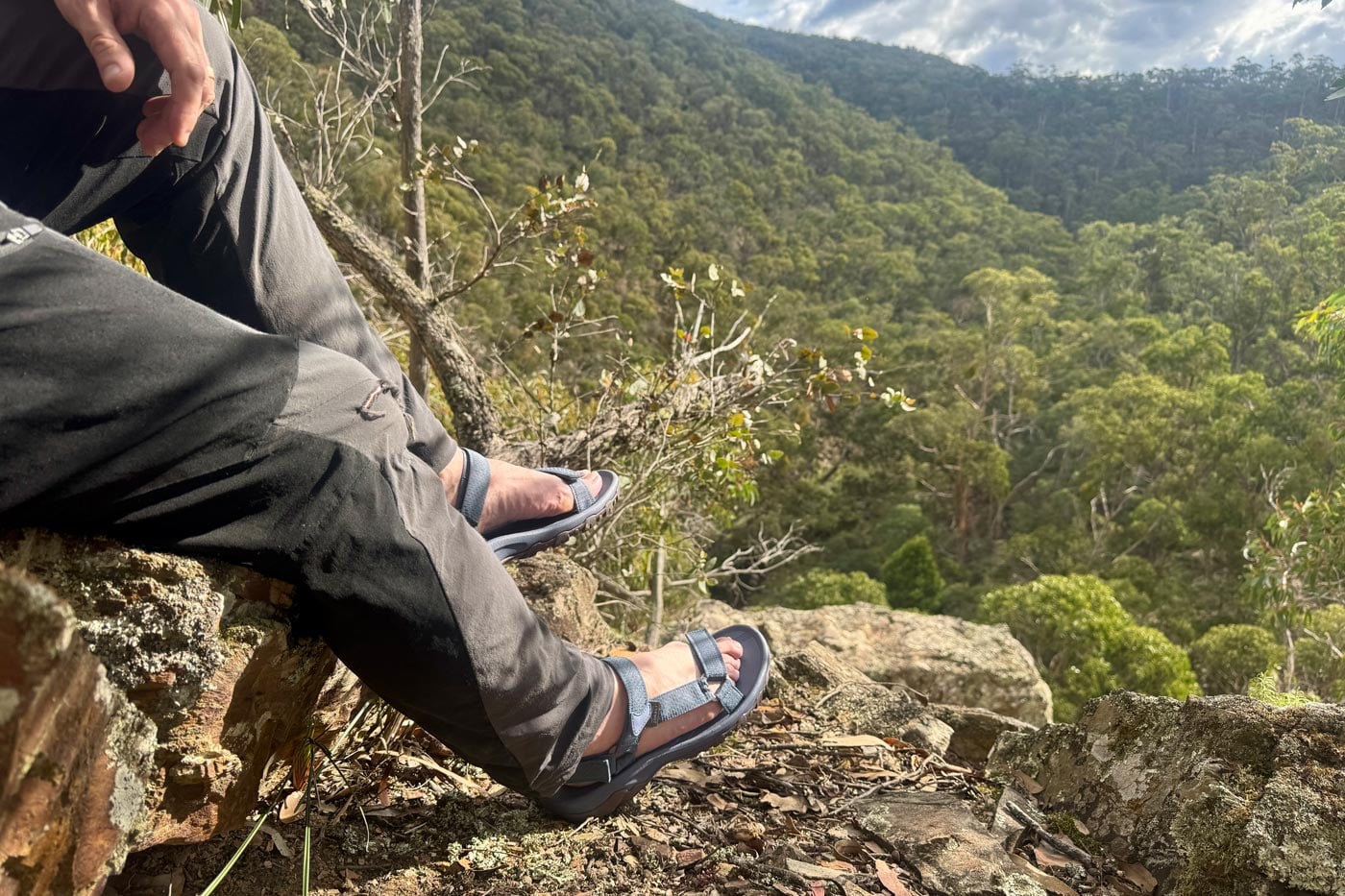 A hiker sitting on a rocky ledge overlooking a lush, forested valley, wearing zorali sandals and dark hiking trousers.