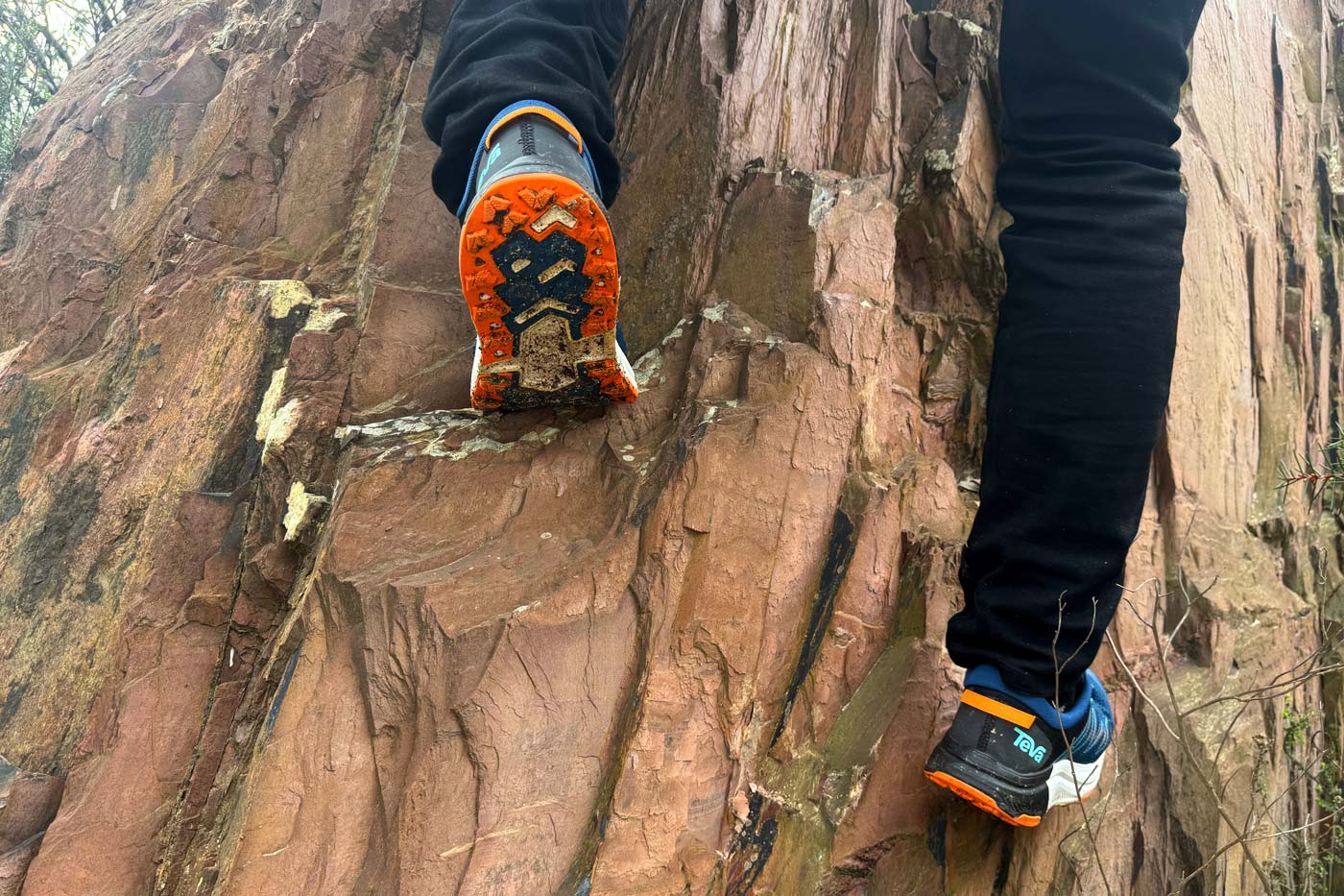 Teva Hurricane Trailsetter Shoe Review Hiker scrambling up a steep rock face wearing teva men's hurricane trailsetter trail shoes, with the orange spider rubber endure outsole clearly visible gripping the rock surface.