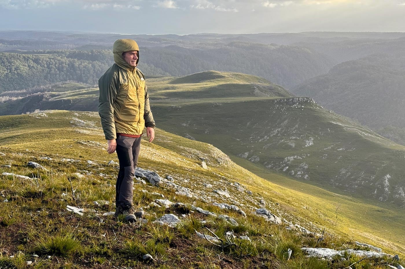 Hiker wearing a ridgelite futurefleece wind jacket stands on an exposed grassy mountain ridge overlooking rolling hills and forested valleys.