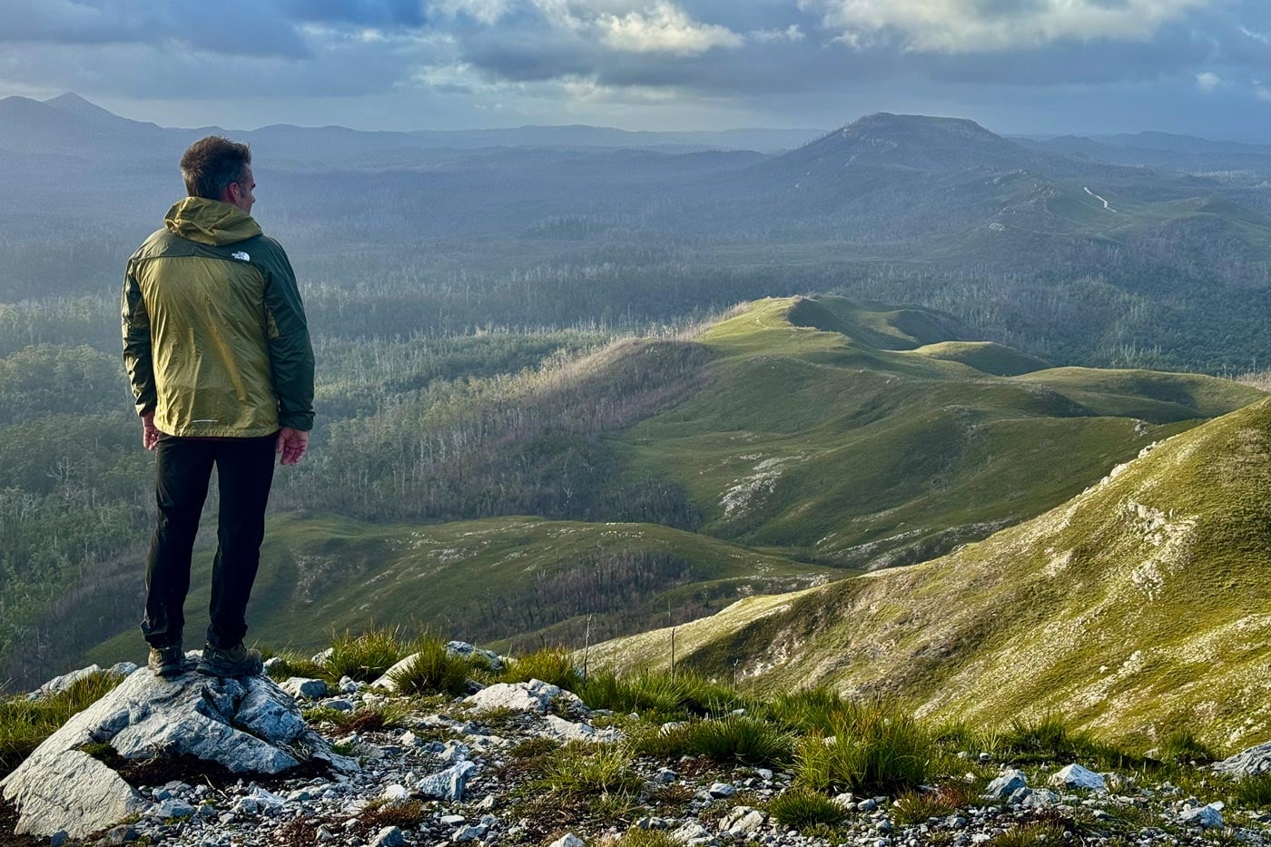 Hiker wearing a ridgelite futurefleece wind jacket stands on a rocky alpine lookout overlooking rolling mountain ridges and forested valleys.