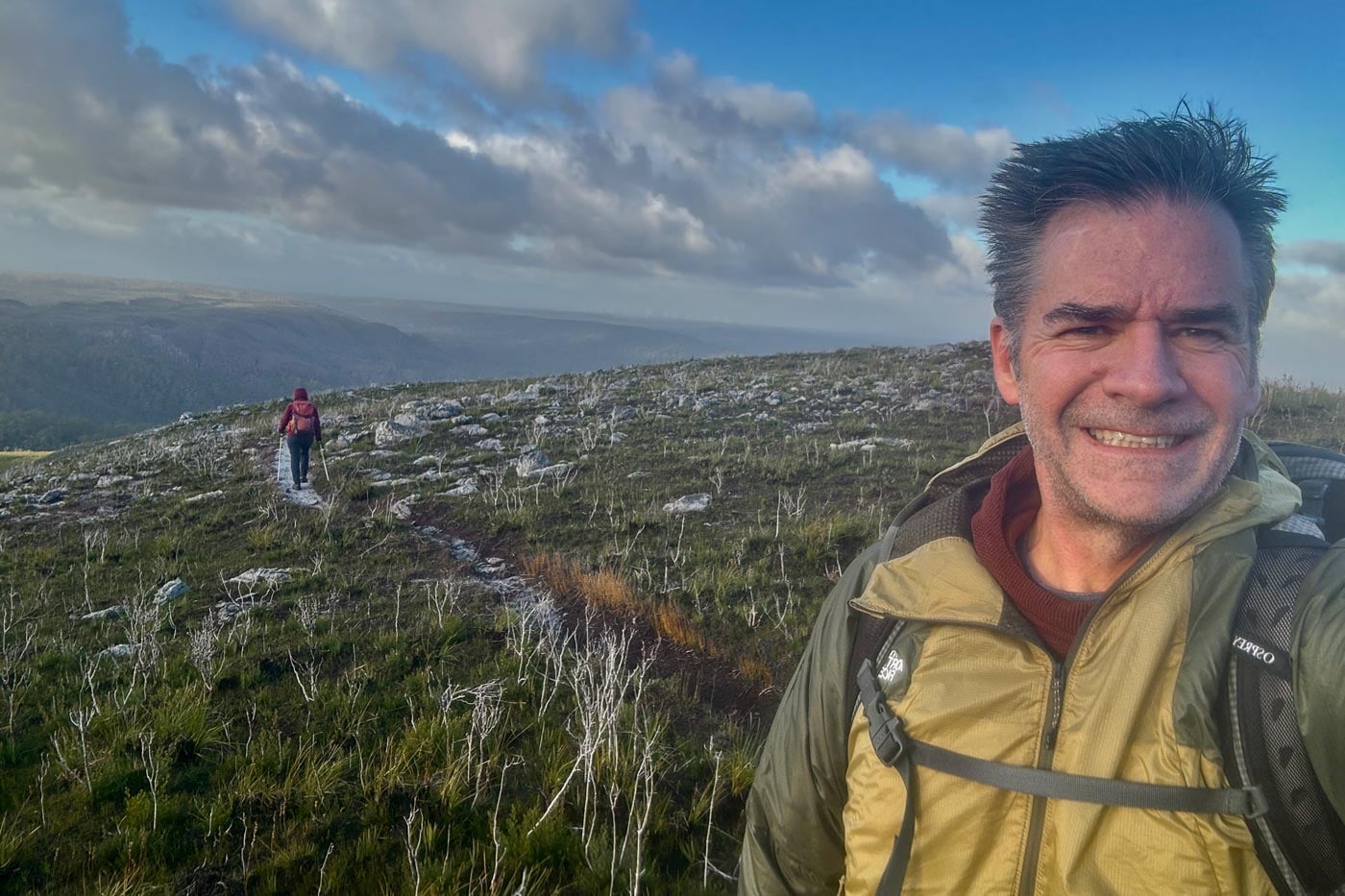 Hiker wearing a ridgelite futurefleece wind jacket with a backpack walks across an exposed alpine plateau under cloudy skies.