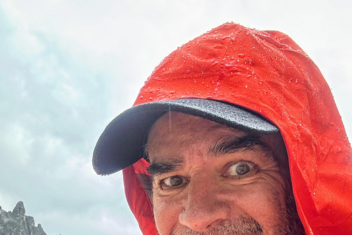 Close-up of hiker wearing the red hood of a waterproof jacket with rain droplets visible on the fabric during wet mountain conditions.