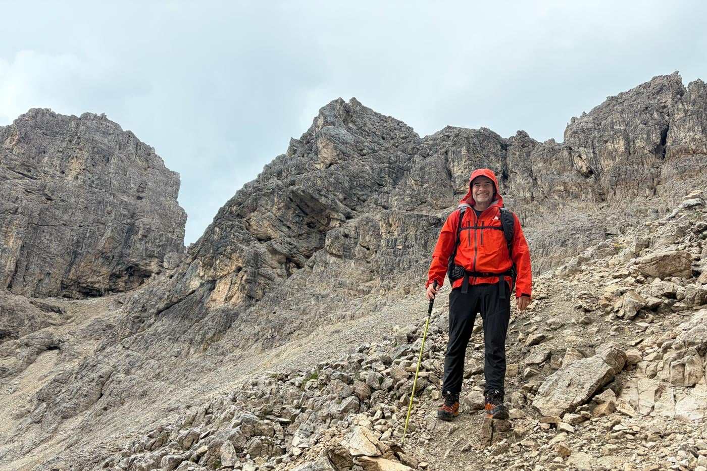 Hiker wearing a red waterproof jacket stands on a rocky alpine trail surrounded by steep dolomite peaks and exposed mountain terrain.