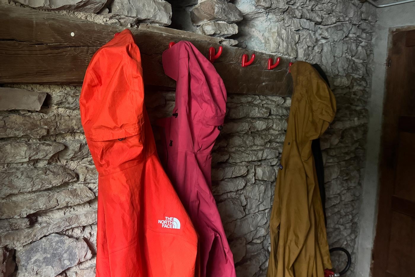 Waterproof jackets hanging on hooks inside a stone mountain hut, showing outdoor gear drying after hiking.
