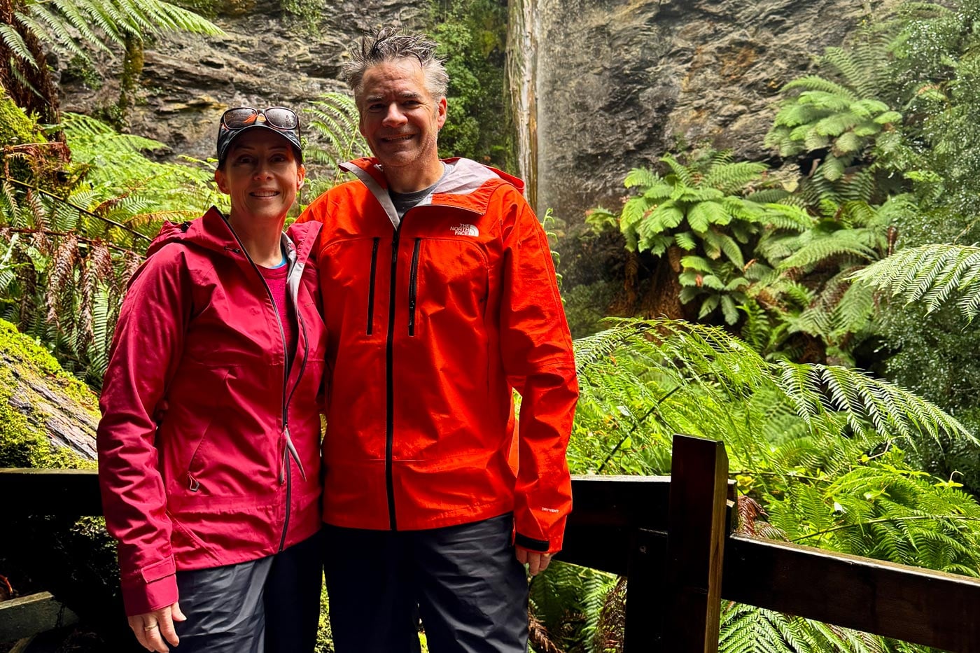 Two hikers wearing waterproof jackets stand on a boardwalk surrounded by lush rainforest and ferns.