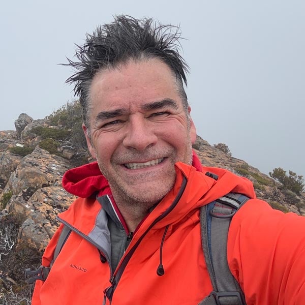 Darren edwards, founder of trail hiking australia in an orange rain jacket smiling for a selfie on a foggy, rocky mountain ridge in tasmania