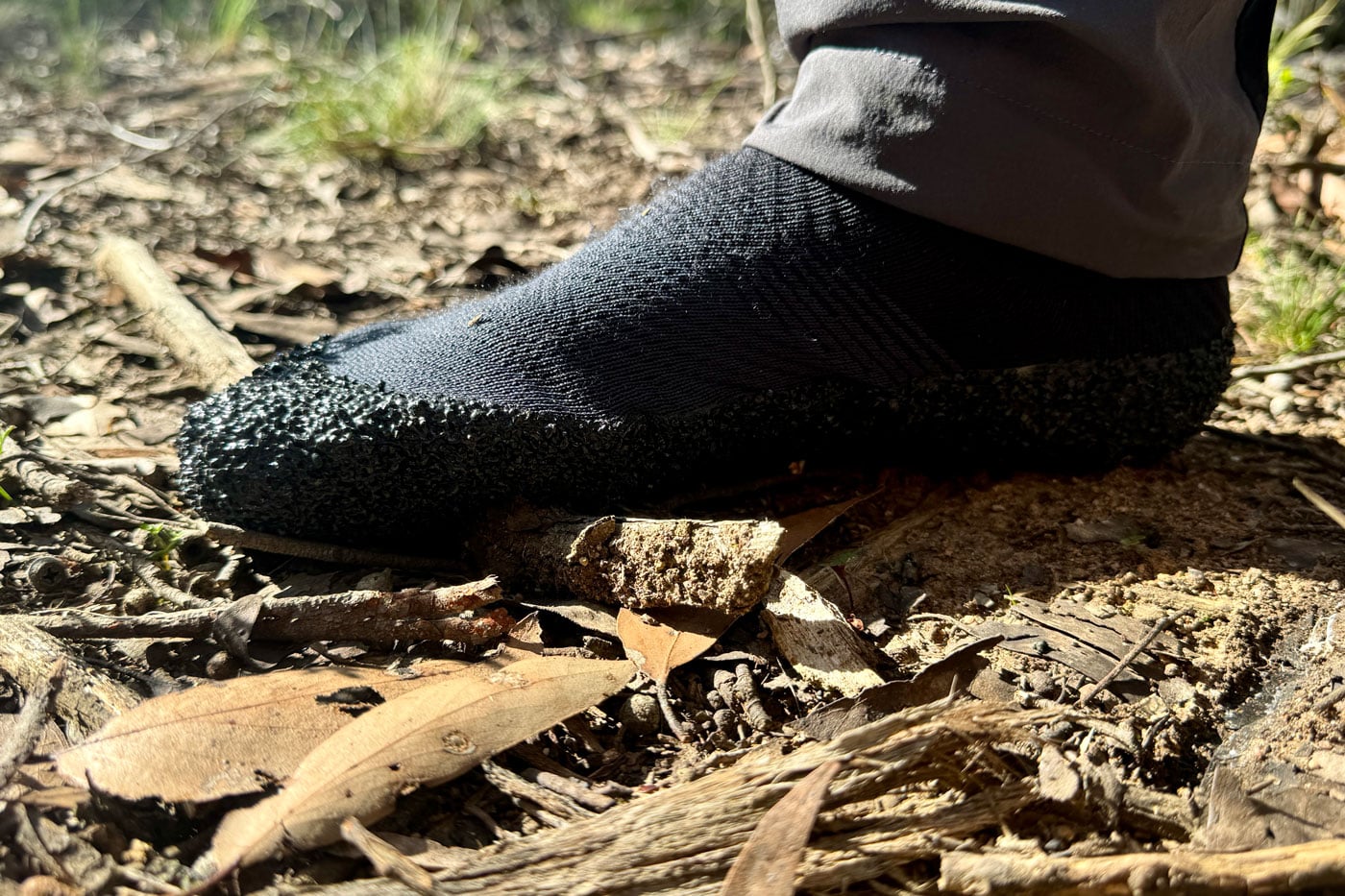 Skinners Comfort 2.0 Sock Shoe Review Close-up of a foot in a black minimalist sock shoe with a grippy rubberised sole, standing on leaf litter and twigs