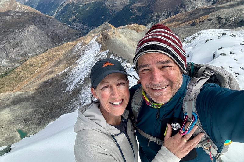 Julie and Darren Edwards Smiling couple, julie and darren edwards, taking a selfie on a snowy mountain ridge, with a valley and peaks in the background.